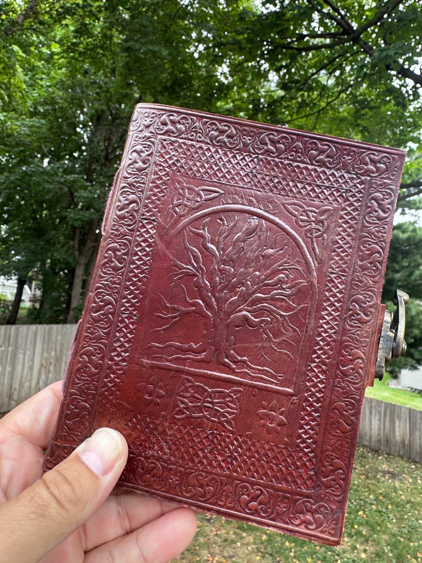 A hand holding an embossed red leather sketchbook beneath green trees